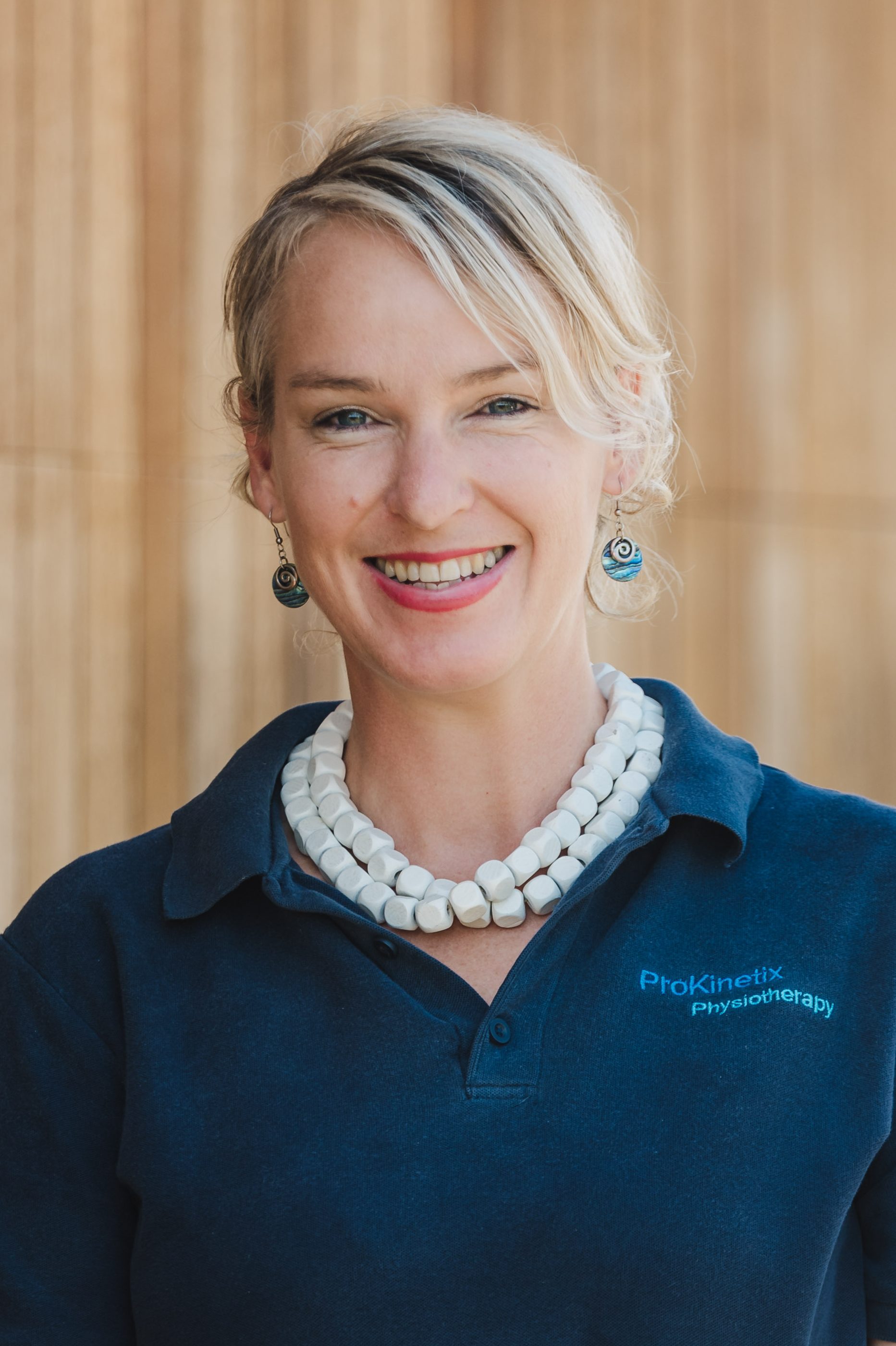 Photo of a blonde woman wearing a white necklace, paua shell earring, and a blue polo shirt that reads, "ProKinetix Physiotherapy". She is smiling.